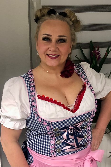 Chris Schlenker im blau-weiß karierten Dirndl mit rosa Schürze, Haarknoten und roter Blüte, lächelnd
