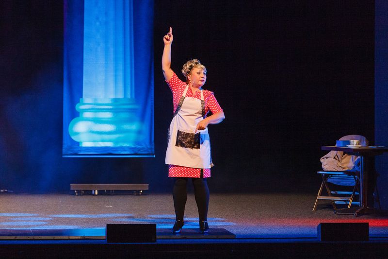 Chris Schlenker als Helga Hahnemann in weißer Schürze über rot gepunktetem Kleid, den Zeigefinger erhoben