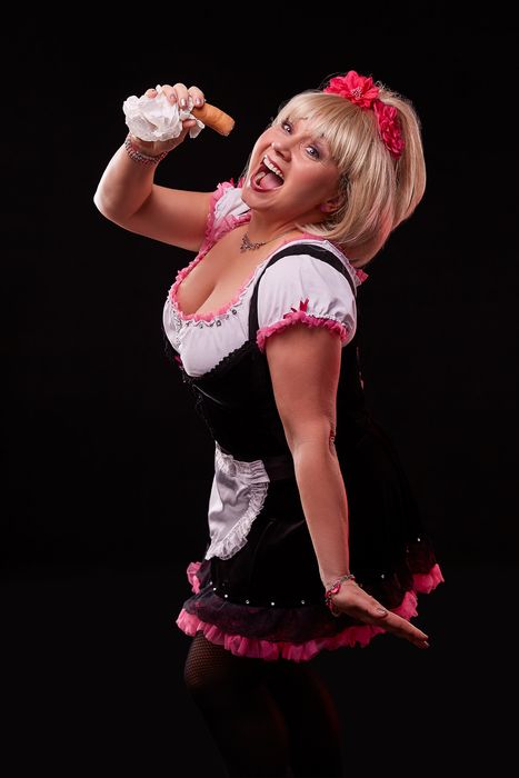 Chris Schlenker als Oberkellnerin Jacquelinchen im schwarz-pinken Dirndl, mit einem Würstchen in der Hand, ausgelassen lachend
